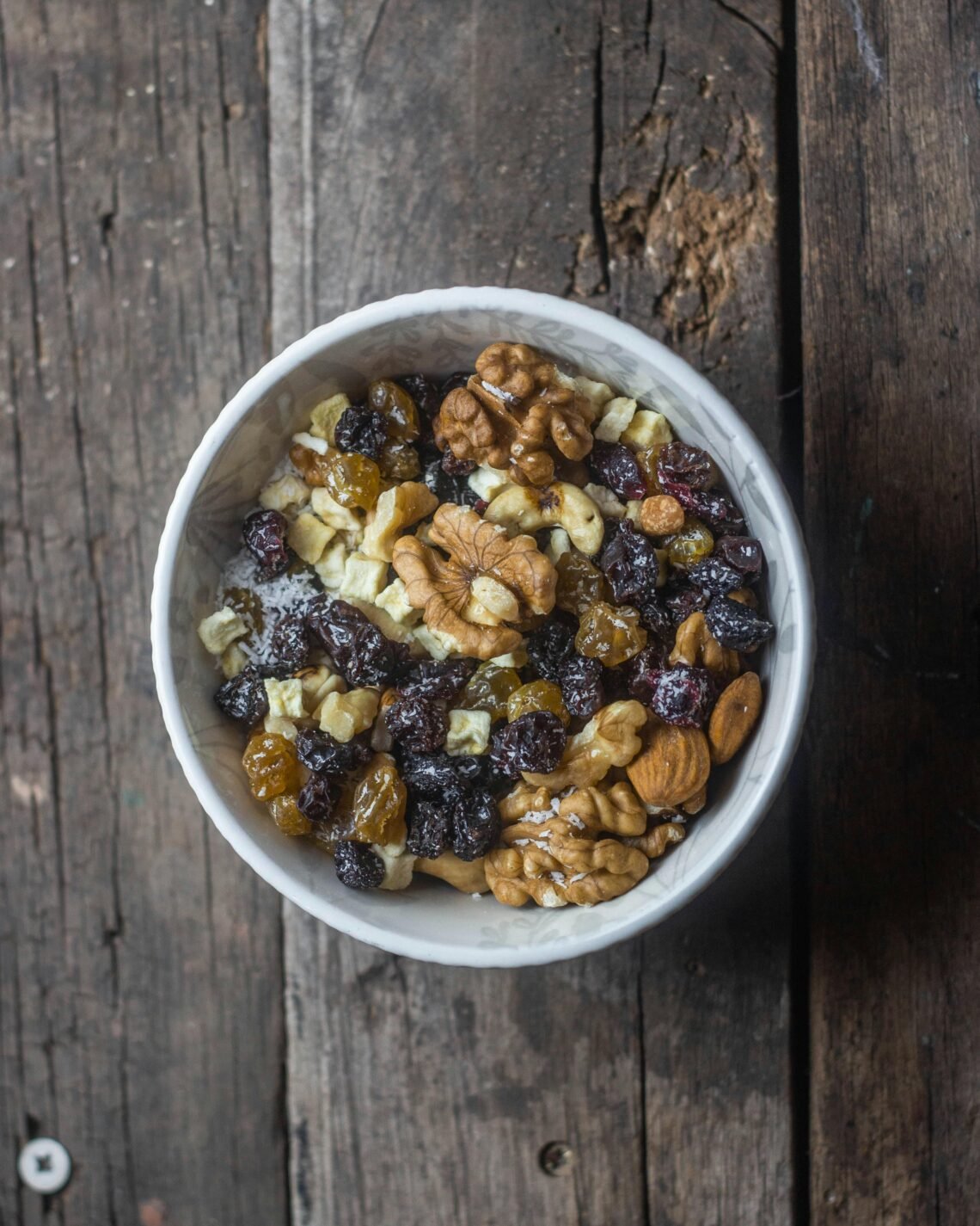 Top view of a bowl with mixed nuts and dried fruits on a rustic wooden table.
