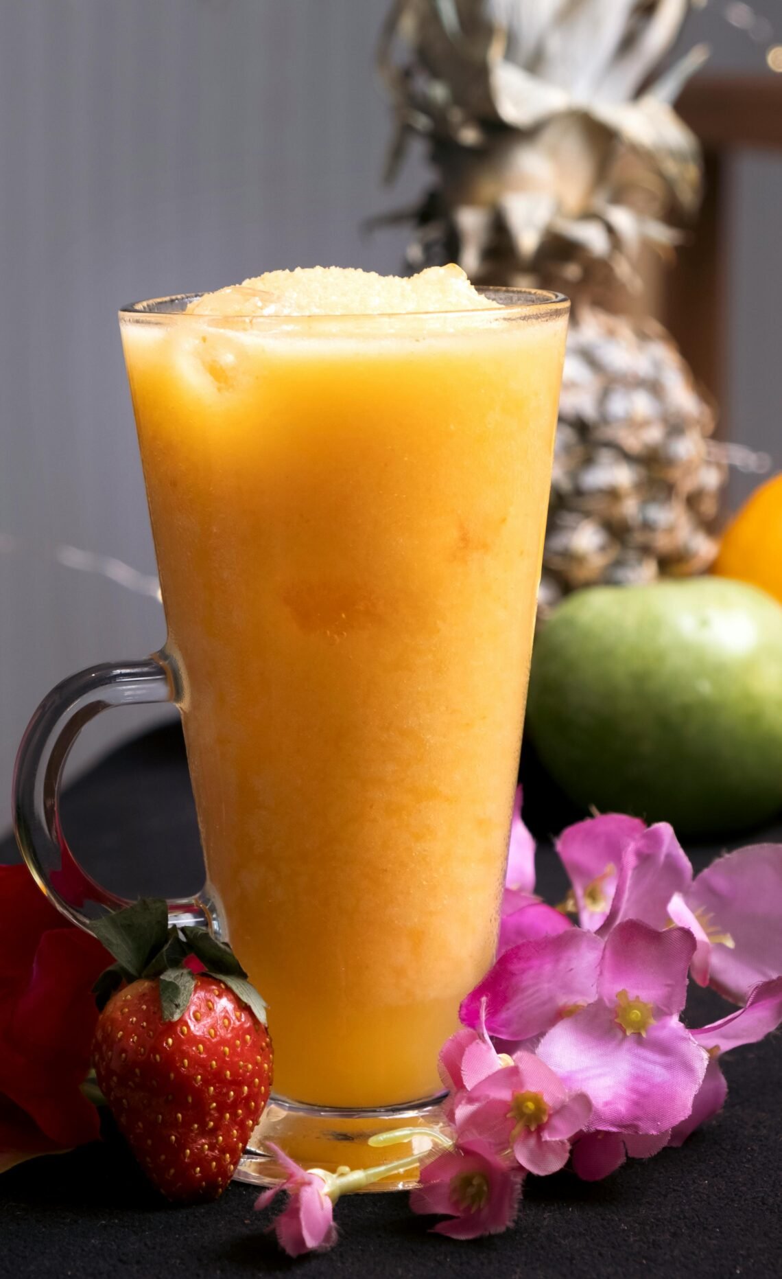 Delicious tropical fruit smoothie in a clear glass mug surrounded by fresh fruits and flowers.