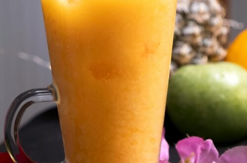 Delicious tropical fruit smoothie in a clear glass mug surrounded by fresh fruits and flowers.