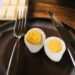 A close-up of a sliced hard boiled egg with a fork and knife on a rustic plate.