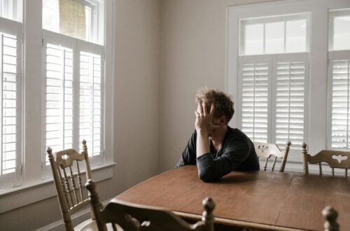 A man sits alone at a table in a bright room, displaying deep contemplation.