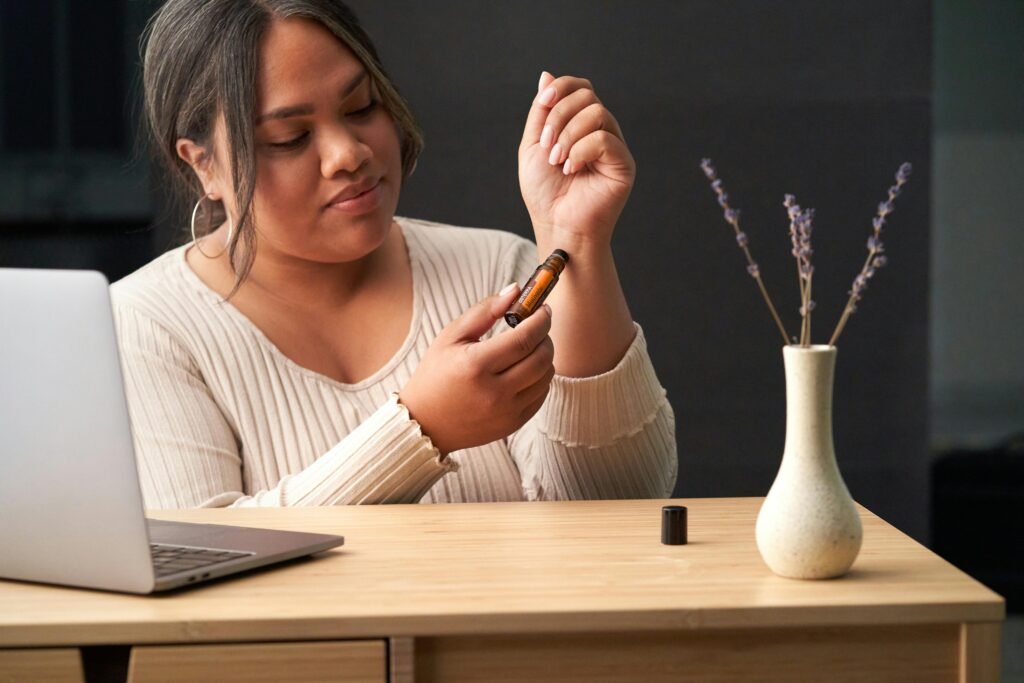 A woman uses a therapeutic essential oil roll-on at a work desk featuring a laptop and vase with flowers.