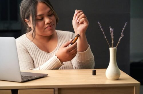 A woman uses a therapeutic essential oil roll-on at a work desk featuring a laptop and vase with flowers.